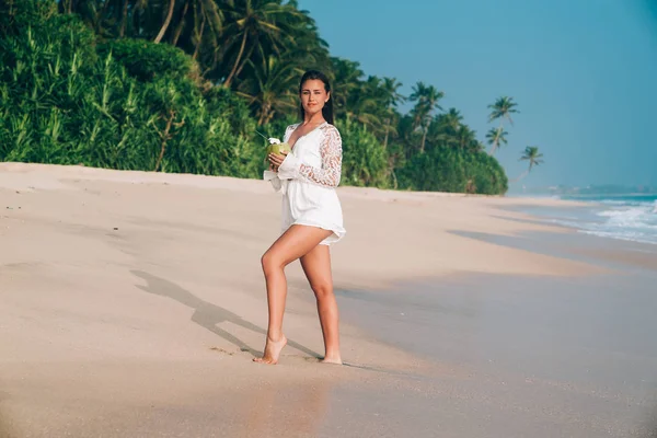 Uma menina bonita, feminina e graciosa voou para descansar, caminha ao longo da praia de areia branca ensolarada, desfruta de descanso, bebe coco fresco . — Fotografia de Stock