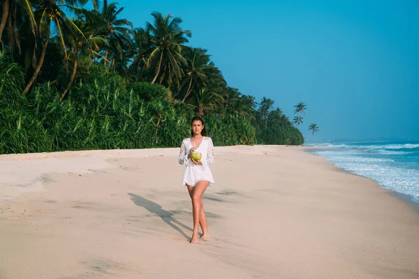 Una bella ragazza gentile in abiti da spiaggia alla moda beve coca e passeggia lungo la riva del mare, ammirando la fitta giungla verde e il mare surf. — Foto Stock