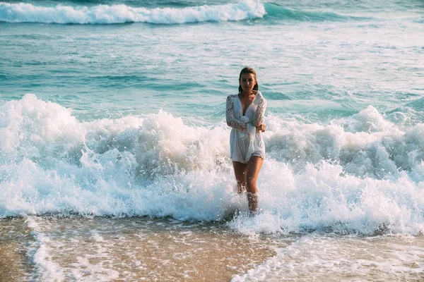 Bronceado hermosa brillante atractiva chica bañada en el mar azul en la ropa, sale del agua, sobre el fondo de las olas y la espuma blanca en un corto blanco en general — Foto de Stock