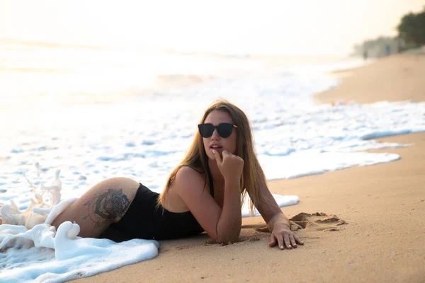 Μια σέξι όμορφη κοπέλα σε ένα κύμινο βρίσκεται στην παραλία, ένα κύμα που καλύπτει τα πόδια της και συνετρίβη κατά τον στρογγυλό κώλο. Έννοια της ανάπαυσης, παραλία, άνθρωποι, ωκεανός — Φωτογραφία Αρχείου