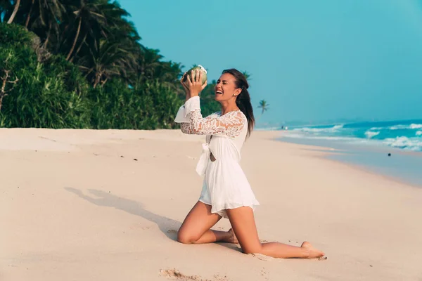 La modelo se ríe de pie en la playa de rodillas, tratando de verter agua del coco en su boca, riendo y divirtiéndose. Una chica trata de beber una pipa de coco sin un tubo . — Foto de Stock
