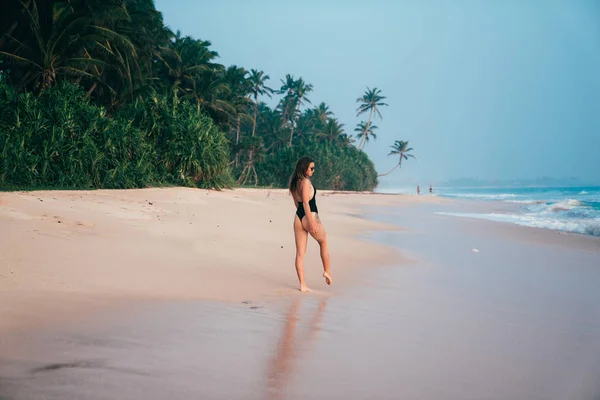 Una giovane ragazza sexy in costume da bagno cammina al tramonto su una spiaggia selvaggia deserta, sullo sfondo di palme, sabbia e onde del mare blu . — Foto Stock