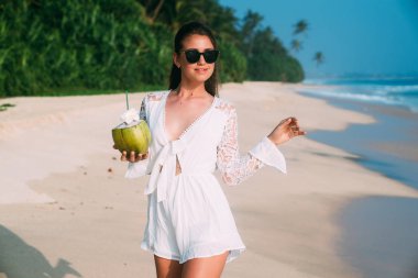 Mutlu esmer denizin üzerinde duruyor, beyaz plaj boyunca yürüyor, Güneş ve okyanus sahiptir, taze Hindistan cevizi suyu içiyor. Kız güneşli şık gözlük ve modaya uygun rahat bir beyaz giymiş