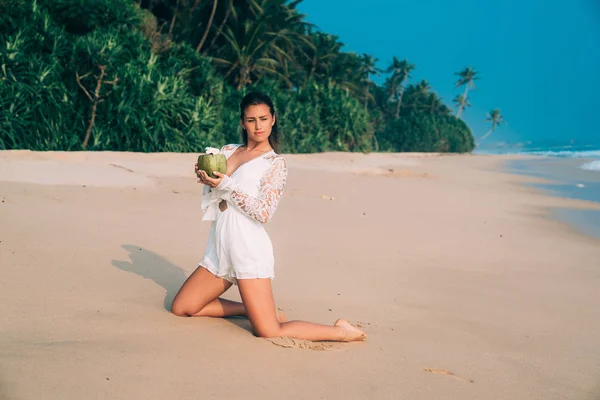 Mladá Evropská žena přišla na tropickém ostrově, klečení na písku a drží čerstvé mladé lahodné kokosové, obdivovat oceán. — Stock fotografie