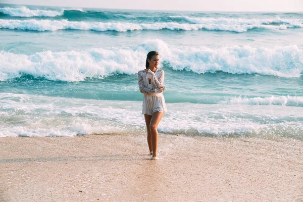 Una hermosa joven europea bronceada y brillante con un traje blanco como la nieve está de pie en la playa sobre un fondo de altas olas azules con espuma blanca, sonriendo y disfrutando de las vacaciones. —  Fotos de Stock