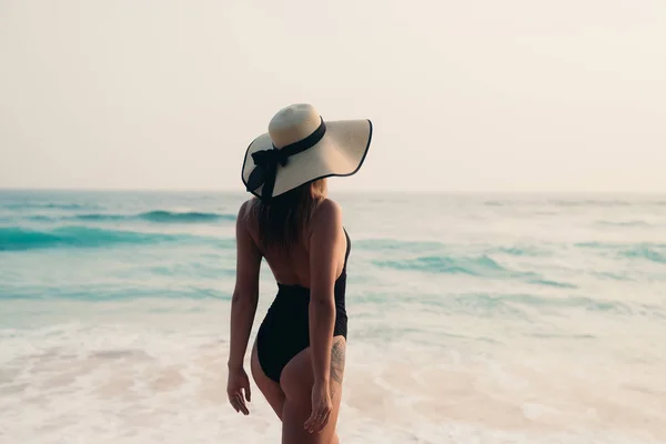 Hermosa chica delgada con un aspecto redondo botín de la parte posterior va contra el mar, vestido con un sombrero caro y bikini. El concepto de relajarse en el mar, la playa, la gente . — Foto de Stock