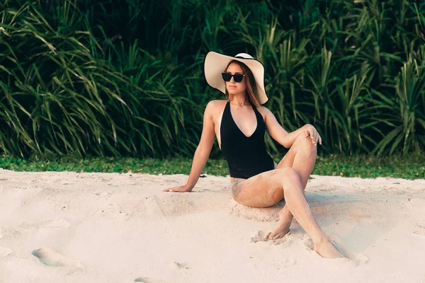 Europeo con morbida pelle liscia poggia sulla spiaggia, prendere il sole e godersi il sole, indossa un elegante costume da bagno nero, cappello e occhiali. Donna d'affari sulla spiaggia — Foto Stock