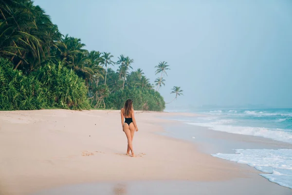 Blick von hinten, ein schlankes Mädchen im schwarzen Badeanzug schlendert am Ufer eines rosafarbenen Strandes vor dem Hintergrund tropischer Dschungel, Palmen und Bäume und blauen Meerwassers. — Stockfoto