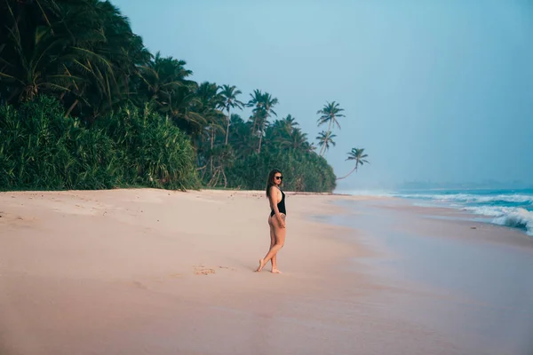 Una hermosa niña pasea a lo largo de una playa tropical con arena rosa, lleva un traje de baño elegante, admira el mar y las palmeras — Foto de Stock