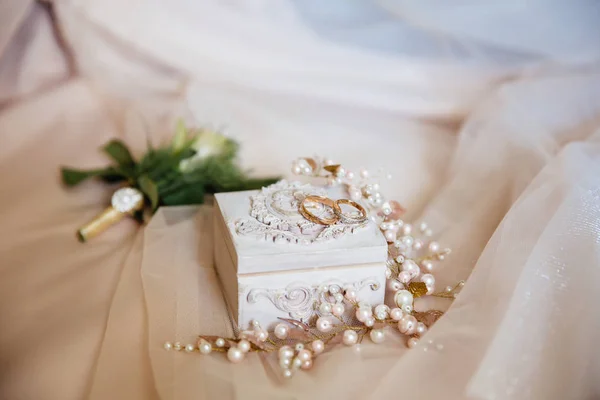 Close-up van bruiloft versieringen en kenmerken. de gouden ringen van de pasgetrouwden liggen op een prachtige witte kistje, een stapel van stengels met witte parel kralen op een zachte beige achtergrond. — Stockfoto