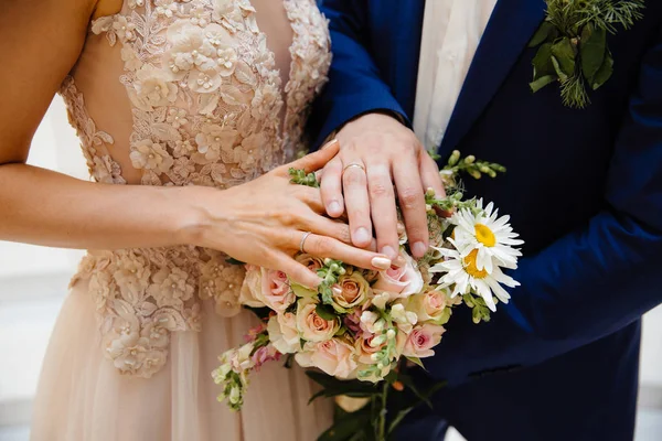Un primer plano de un marco recortado de la novia y el novio de pie lado a lado y tocando su ramo de bodas, mostrando sus anillos de boda en las patas . — Foto de Stock