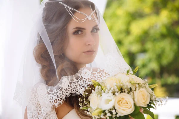 美しい若い花嫁がきれいなは、皮膚、クローズ アップ。ウェディング ベールを通して女の子の顔。白バラからの花嫁の花束. — ストック写真