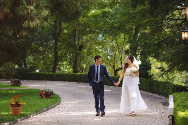 Закохані щасливо і щасливо ходять по алеї в зеленому щільному парку, спілкуються і сміються в день весілля, тримаючи руки . — стокове фото