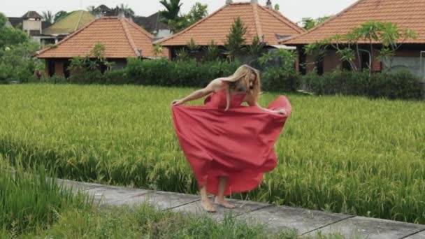 Una mujer con una figura proyectiva baila sola en el campo de arroz, toma su falda larga y falda de un vestido brillante, muestra sus piernas, salta y corre en el marco, hace un cordel vertical en un lento — Vídeo de stock