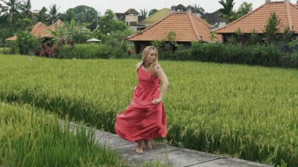 Танцовщица носит длинное красное платье, ее светлые волосы расцветают, а губы окрашены яркой помадой, она танцует и поворачивается на рисовом поле, сохраняет ее развивающуюся юбку, эмоционально — стоковое видео