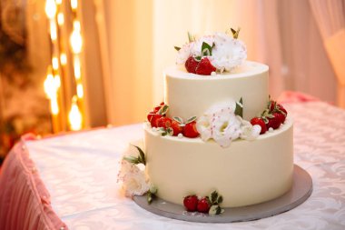 Düğün üç katlı beyaz kremalı pasta tablodur. Tatlı olgun kırmızı çilek çilek ile dekore edilmiştir ve beyaz çiçekler yaşamak. Stil düğün düğün pasta dekorasyonu için fikirler.