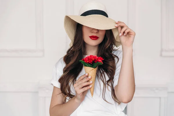Fiatal gyönyörű nő vegán portré, nagy mezők szalma kalap viselése, és amely a szeme, vörös ajkak. A lány azt hiszi, hogy édességek káros az a szám, így ő már piros virágok a kürt. — Stock Fotó