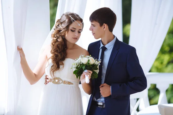Die schöne Braut trägt ein Kleid im griechischen Stil, ihr Haar ist mit Perlen verziert, sie hält einen klassischen Hochzeitsstrauß weißer Rosen in der Hand und blickt ihren schlanken, großen Mann in einem blauen Geschäft an — Stockfoto