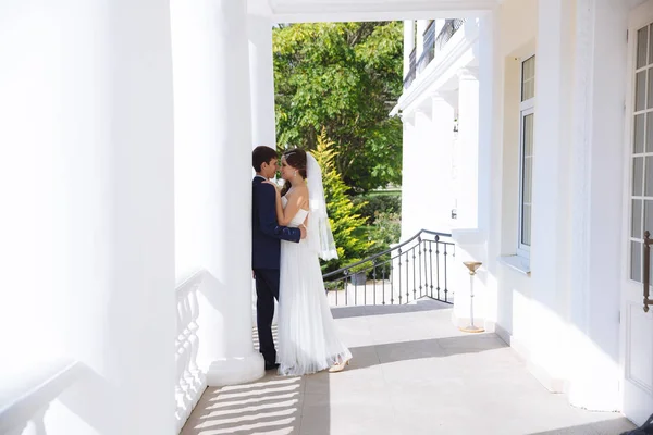 La bella sposa è vestita in un abito in stile greco, i suoi capelli sono decorati con perline, sta tenendo un classico bouquet da sposa di rose bianche, guardando il suo snello marito alto in un business blu — Foto Stock