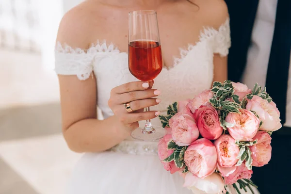 Крупним планом обрізана рамка нареченої в білій сукні тримає в руці весільний букет з рожевих півоній, келих рожевого шампанського, одягнений в плоске заручини золоте кільце. Манікюр нареченої — стокове фото