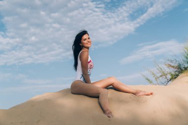 Açık bir kar-beyaz gülümseme ile neşeli çekici kız sahilde alt görünümü dinleniyor, model mavi gökyüzüne karşı kumlu bir tepe üzerinde oturuyor. Kız tasasız bir tatil sahiptir.