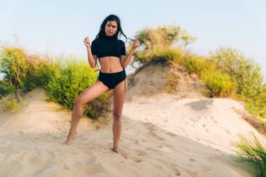 Uzun kıvırcık saçlı çekici genç kız şakacı kendini bağları bir mayo üzerinde üzerinde vahşi çalı zemin karşı beyaz bir plajda poz çeker, vahşi bir plajda bir Amazon gibi geliyor.