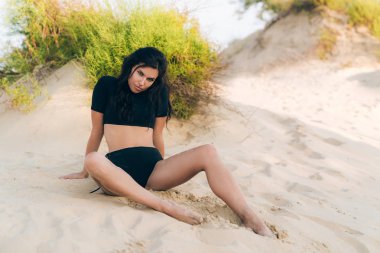 Baştan çıkarıcı kız bir şık mayo bir ıssız vahşi sahilde poz sıcak kum üzerinde yatıyor, seductively çerçeve içine görünüyor.