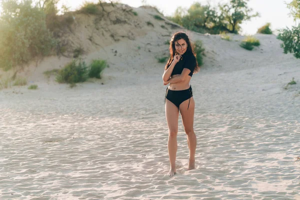 Una chica bronceada atractiva camina a lo largo de una playa vacía con arena blanca, lleva un traje de baño negro separado de moda, tiene el pelo oscuro rizado, sonríe. Concepto de descanso, viajes, personas . — Foto de Stock