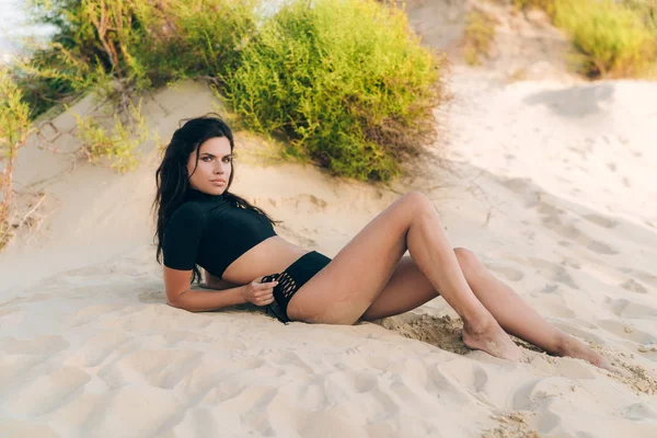 Smal elegant modell med europeiskt utseende innebär för reklam baddräkter på en öde strand. Flickan är klädd i en separat sluten surfer baddräkt, liggande på sanden. — Stockfoto