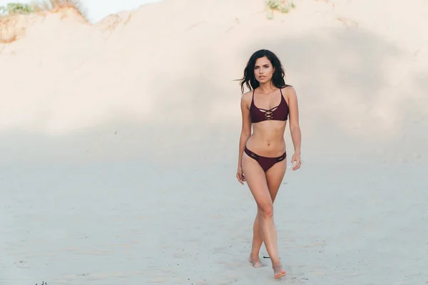 Una chica atractiva joven tiene una figura esbelta inteligente caminando alrededor de una playa rocosa desierta sola, con un elegante bikini de traje de baño de color burdeos, tiene el pelo rizado y un aspecto penetrante . —  Fotos de Stock