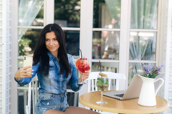 Atractiva morena de pelo largo, de aspecto europeo, elegantemente vestida con jeans, hace selfie en un smartphone con un vaso de zumo de pomelo . — Foto de Stock