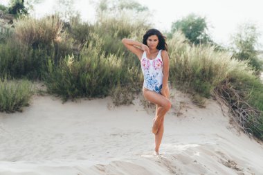 İnce bir model şık bir mayo beyaz kum üzerinde poz dikkatle içine belgili tanımlık çerçevelemek, tek ayak üzerinde duruyor görünüyor, ikinci cinsel diz bükülmüş, karanlık uzun saçları ile Rüzgar çalış.