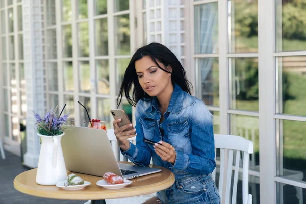 Eine attraktive junge Frau bestellt online, benutzt dazu Telefon, Laptop und ihre Bankkarte, bezahlt ihren Einkauf über das Internet. Gadgets, Technologie, Menschen. — Stockfoto