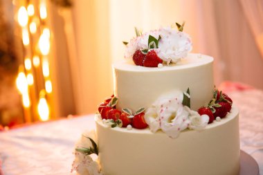 Düğün üç katlı beyaz kremalı pasta tablodur. Tatlı olgun kırmızı çilek çilek ile dekore edilmiştir ve beyaz çiçekler yaşamak. Stil düğün düğün pasta dekorasyonu için fikirler.