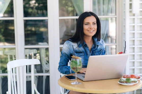 Ein schönes junges Mädchen beschäftigt sich mit irgendetwas auf ihrem Laptop, lächelt, arbeitet an irgendetwas. das Model sitzt in einem Café, trinkt Cocktails, nennt sich Kellner, will etwas anderes bestellen — Stockfoto