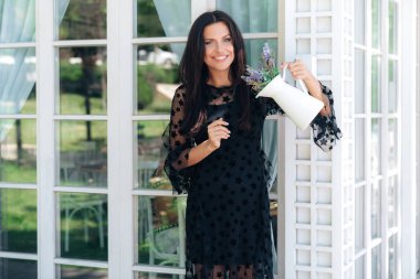 Çekici parlayan kız Avrupa bir görünüme sahip, uzun kavisli bir elbise giyer, şık kenarındaki çiçek dolu vazolar onun elinde, gülümsüyor tutar. Bir ülkenin ev coziness kavramı.