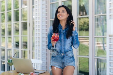Gülümseyen ince güzel kadın şık denim giysileri giymiş, güzel uzun bacakları, lezzetli berry kokteyl içer, vernadzhe bir kafede arkadaşlarıyla tanışmak mutlu zaman harcıyor.