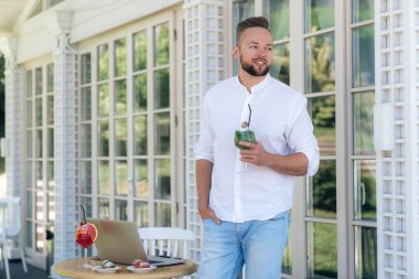 Şık ve çekici bir adam bir kafede standları, nane bir kadeh kokteyl şık beyaz bir gömlek ve mavi kot pantolon giymiş elinde tutan, bir Avrupa görünümü ve şık bir saç kesimi var..