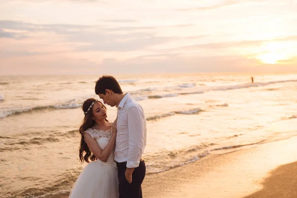 Una bella coppia di sposi novelli, gli sposi che camminano sulla spiaggia. Splendido tramonto e cielo. Abiti da sposa, un abito bianco di lusso per una ragazza. Concetto di famiglia, luna di miele . — Foto Stock