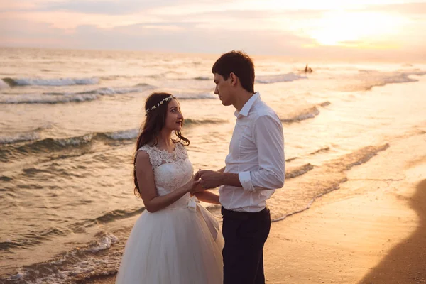 Una bella coppia di sposi novelli, gli sposi che camminano sulla spiaggia. Splendido tramonto e cielo. Abiti da sposa, un abito bianco di lusso per una ragazza. Concetto di famiglia, luna di miele . — Foto Stock