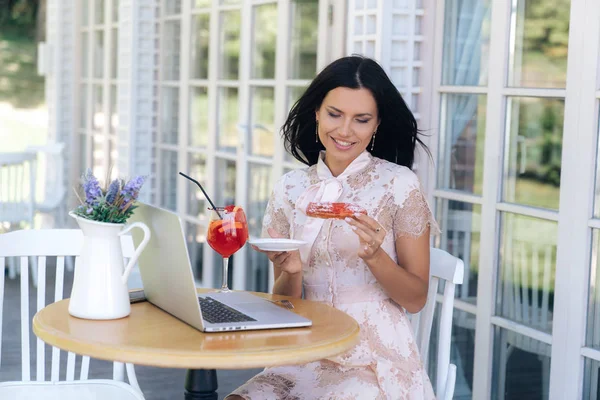 Sonriente jovencita está esperando a sus amigos en un café, se ve algo en su computadora portátil, quiere comer su eclair, está feliz con un buen día y su almuerzo. La modelo está vestida con un delicado vestido de encaje . — Foto de Stock