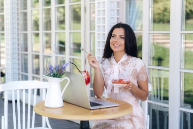 Gülümseyen genç kız Avrupa bir görünüme sahip ve pasta dükkana gitti, sokakta küçük bir masada oturdu, iş arkadaşları, gülüyor için bekledi bir tatlı sipariş, onu laptop verdi.