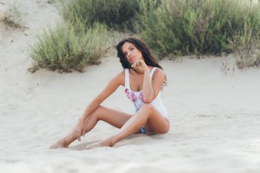 Avrupalı seksi bir Bayan plajda, sıcak kum, oturur onun moda mayoyla poz aittir, Rüzgar uzun siyah saçları geliştirir. Seyahat, turizm, rekreasyon, insanlar kavramı.