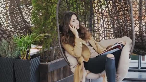 Ein schönes Mädchen in Pullover und Mantel sitzt auf einem Stuhl in einem Café und telefoniert emotional. Frau löst geschäftliche Probleme und hält eine Zeitschrift — Stockvideo