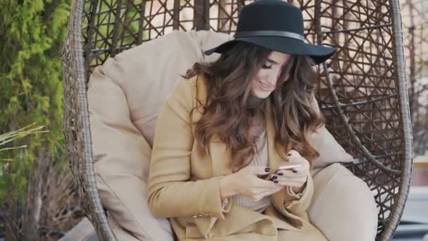 A beautiful young woman wearing a coat and a black hat on her head is sitting in a chair in a cafe and typing sms in the phone. Woman solves business issues and holds smartphone — Stock Video