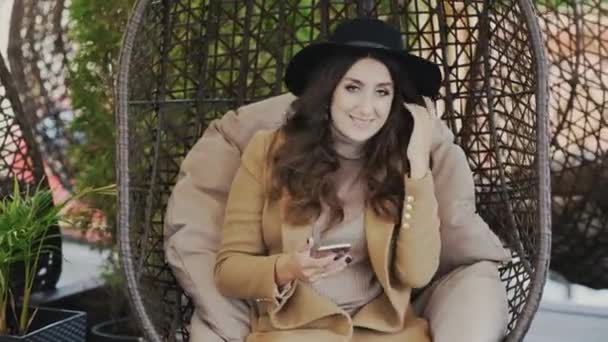 Una hermosa joven sonriente con un abrigo y un sombrero negro en la cabeza está sentada en una silla en un café y escribiendo sms en el teléfono. Mujer resuelve problemas de negocio y sostiene el teléfono inteligente . — Vídeos de Stock