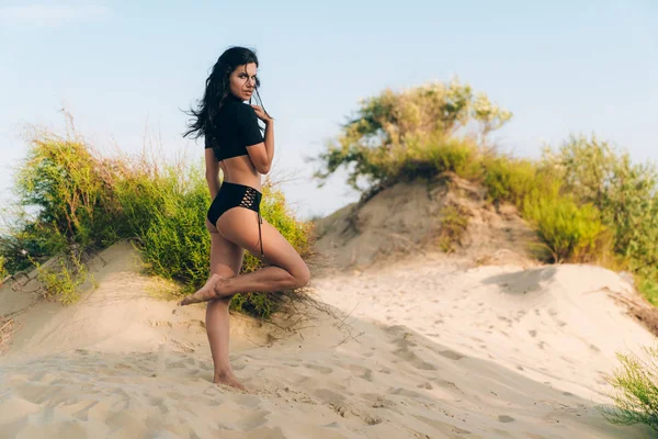 Verleidelijk jong meisje met donkere krullend haar poseren op een wit strand tegen de achtergrond van wilde struiken, tonen van haar slanke lange benen, heeft een gladde warme huid, het dragen van een aparte zwarte zwembroek — Stockfoto