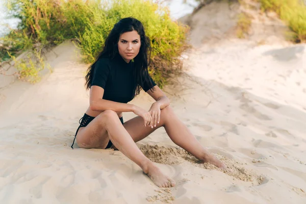 Gebruinde brunette Europese poseren op een wit strand, zittend op het zand, ozhyhayet na het zwemmen in de Oceaan, heeft een gladde warme huid, vernauwde ogen, een sexy look. — Stockfoto