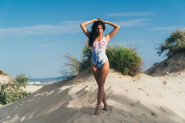 A beautiful sexy European woman with tanned skin walks along the white sandy beach, wears a trendy indoor swimsuit, poses against the background of the sea. The concept of leisure, travel, people.