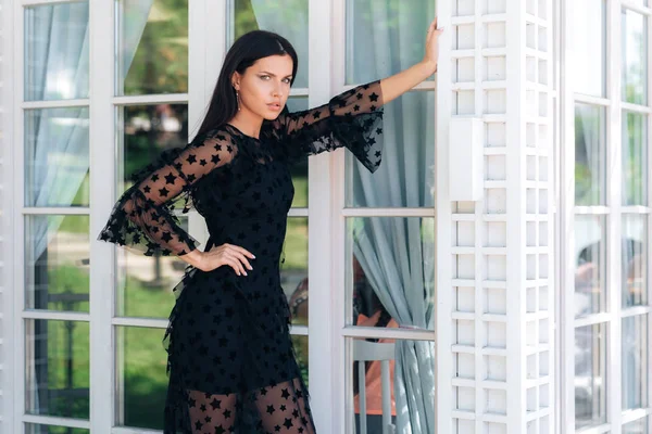 Стильная высокая брюнетка с красивыми длинными ногами позирует в черном платье снаружи у стеклянной стены. Привлекательная девушка снимается для модного журнала . — стоковое фото